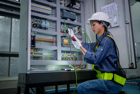 A female technician wearing a hard hat and safety vest works on an open electrical control panel, testing wiring with a multimeter. The image shows industrial electrical infrastructure, technical skills, and hands-on maintenance work in a controlled indoor facility.