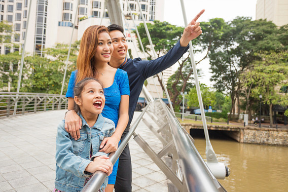 A family of three stands on a pedestrian bridge in a city park, with parents and a young child looking ahead. The father points into the distance while the mother and child smile. The scene depicts an urban lifestyle and family life, with modern buildings, greenery, and a river in the background.